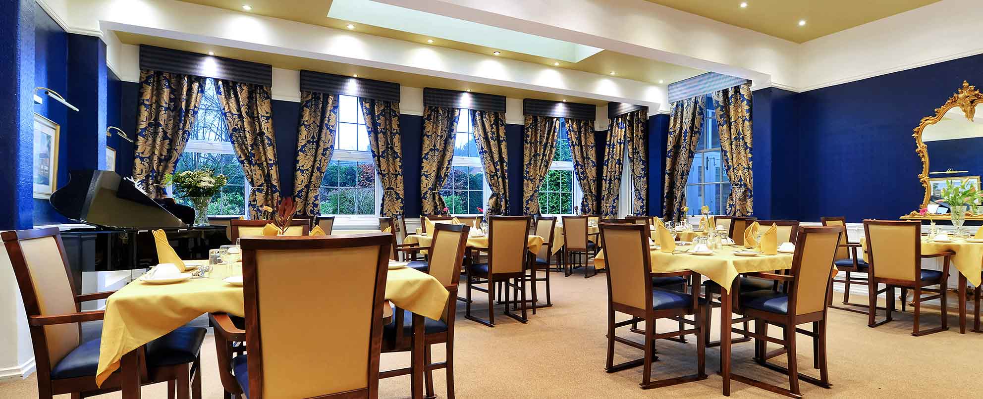 An elegant dining room with multiple set tables, beige chairs, blue walls, and large windows with patterned curtains.