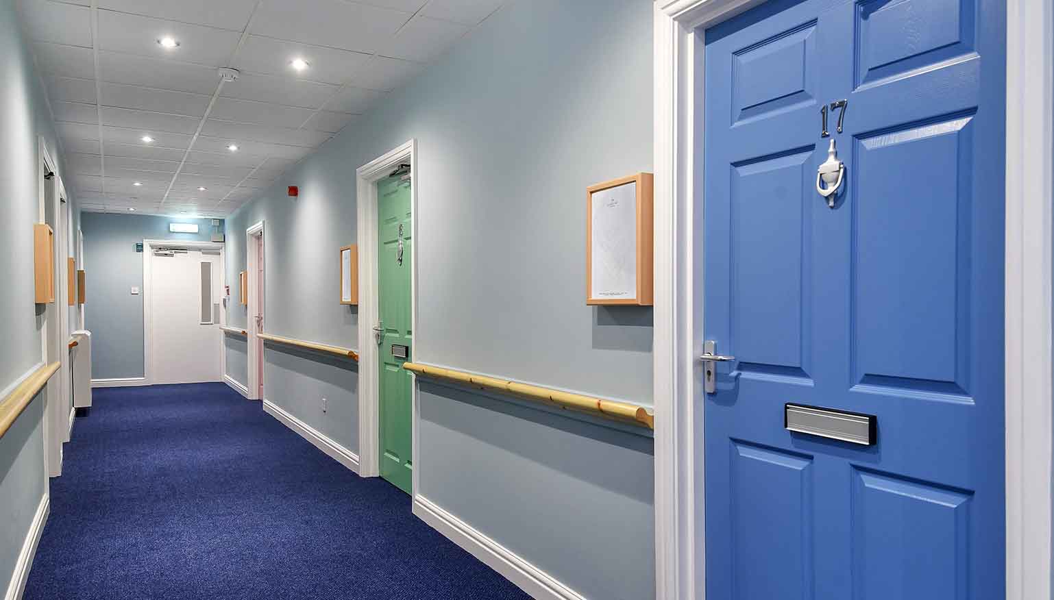 A corridor with blue carpeting and walls painted in light blue and white. There are multiple closed doors with different colours, and a handrail running along one side.