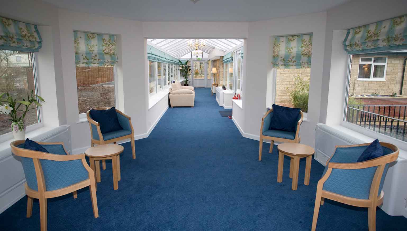 A spacious corridor with blue carpet, white walls, multiple chairs with blue cushions, and large windows allowing ample natural light