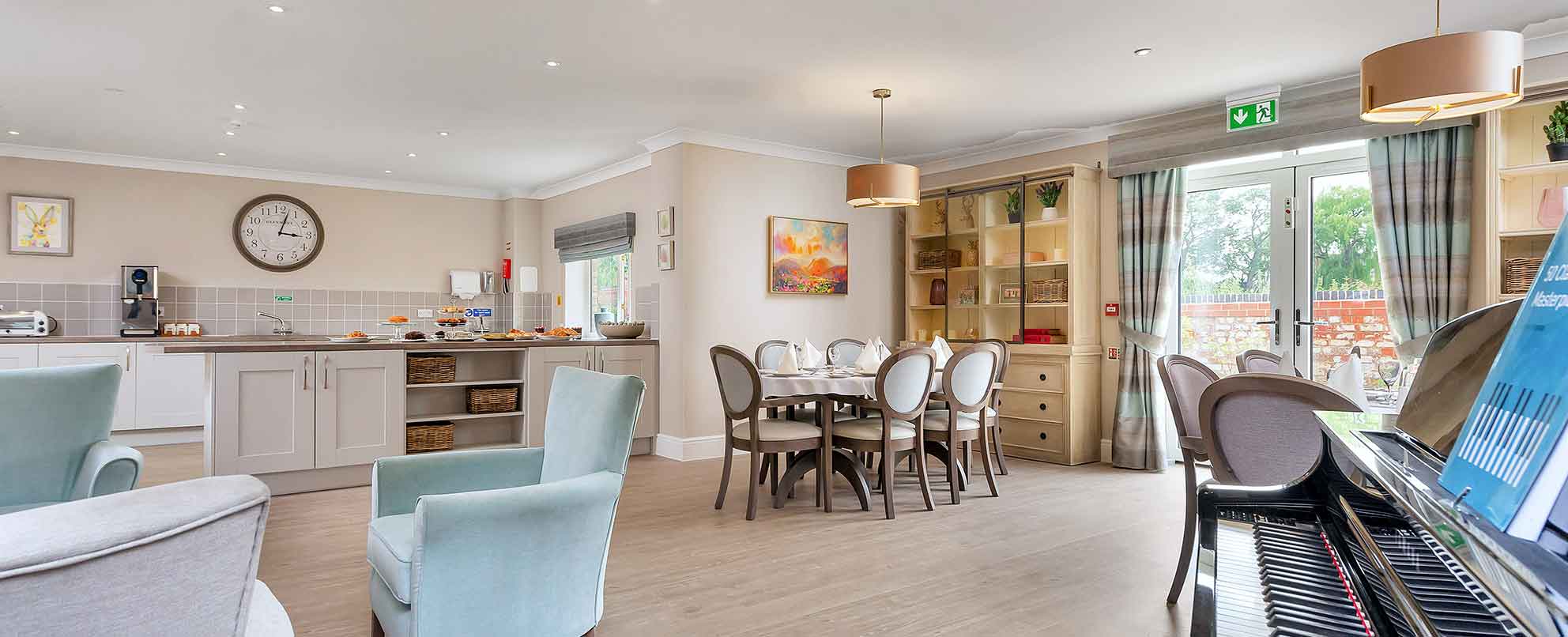 A kitchenette stocked with cakes along with laid dining table, blue lounge chairs and a piano