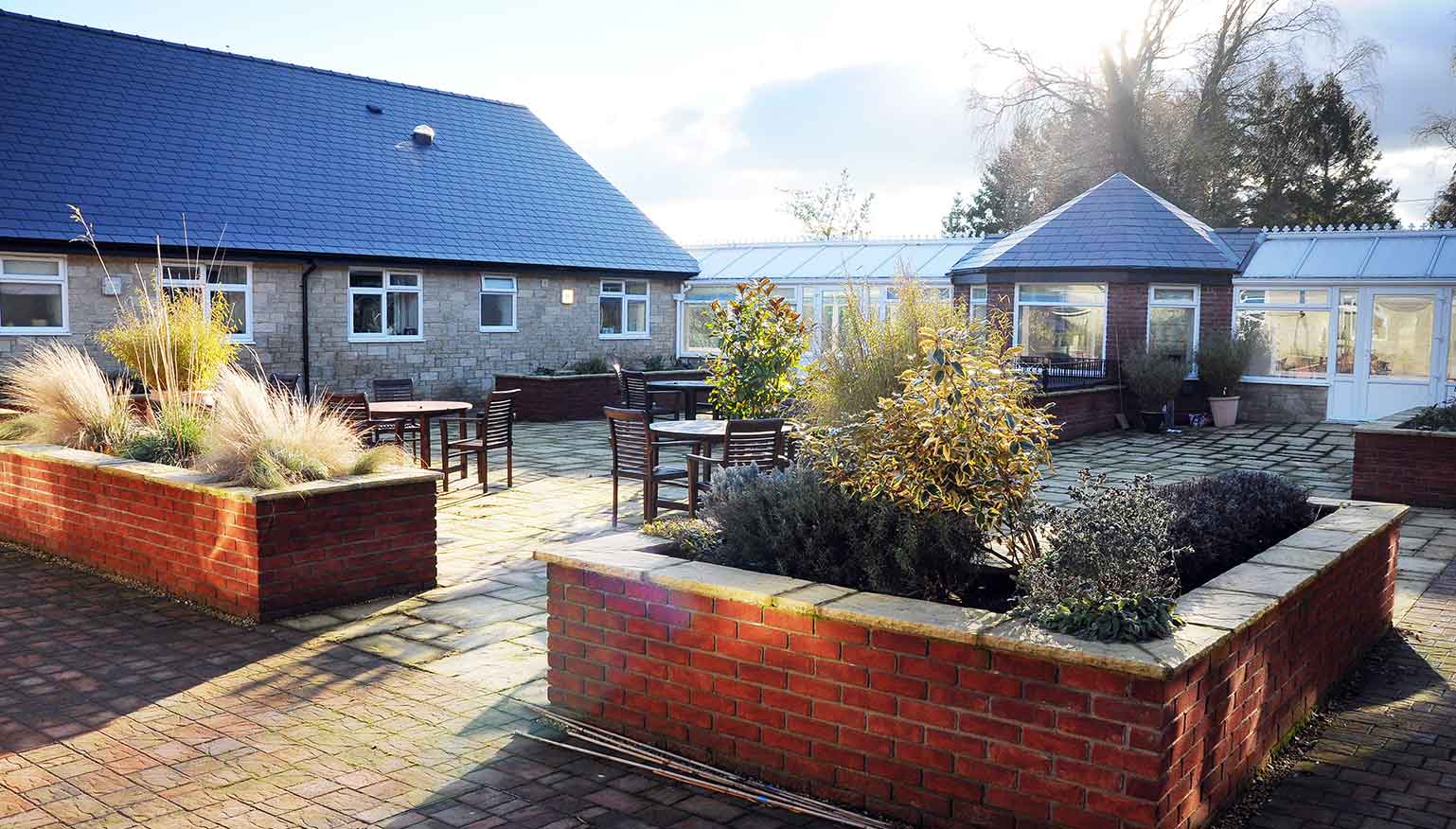 A courtyard with brick planters containing shrubs, a few outdoor tables and chairs, and buildings with blue roofs under a clear sky