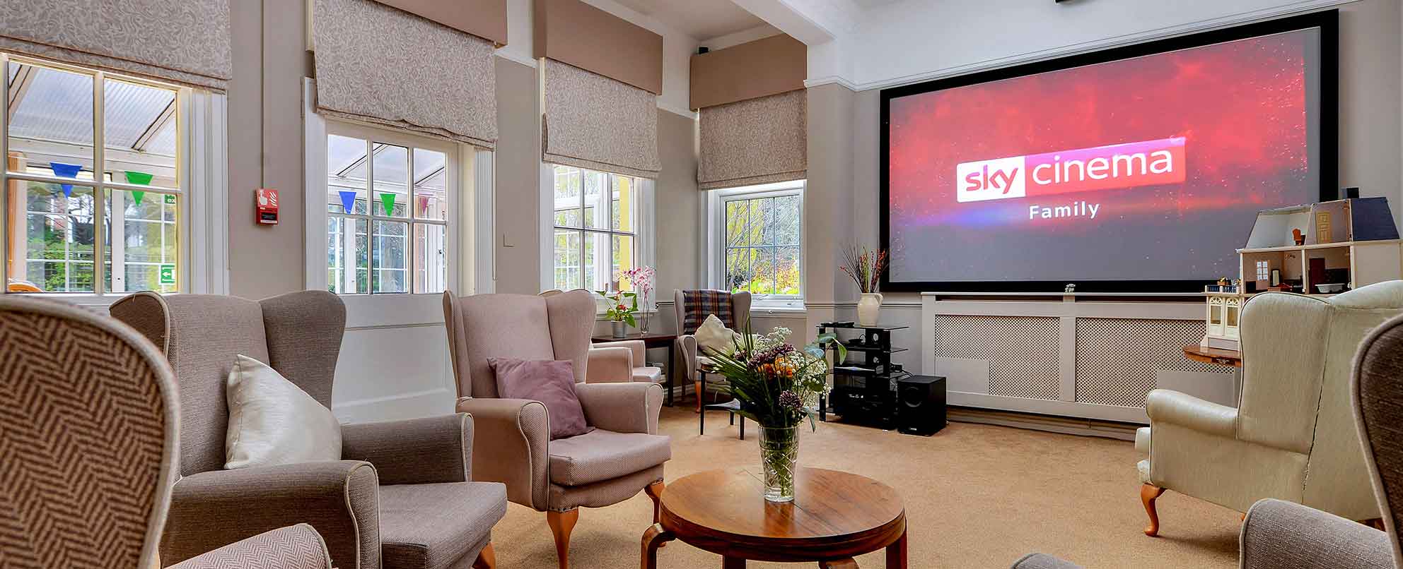 Cinema room with multiple beige armchairs facing a large screen displaying the “Sky Cinema Family” logo. The room has windows on one side, cream walls, and a patterned carpet.