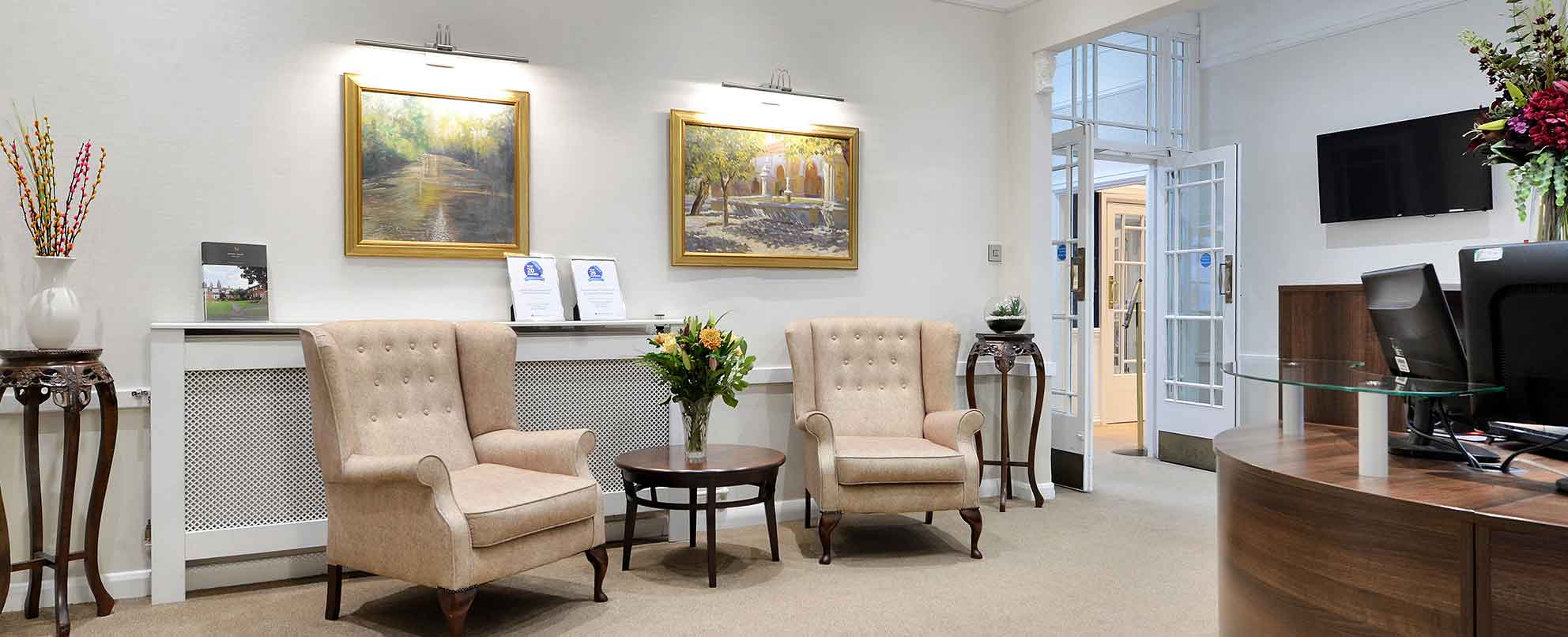 Well-lit reception area with wooden desk, two beige upholstered chairs, a small round table in between, and framed paintings on the wall.