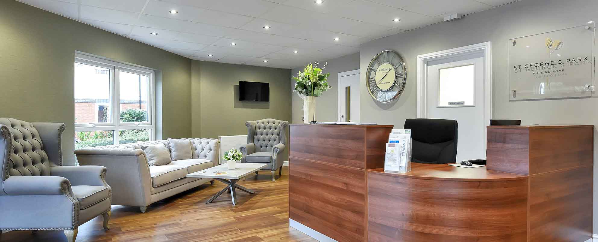 A modern reception area with a wooden desk, grey armchairs, a sofa, and a wall-mounted television