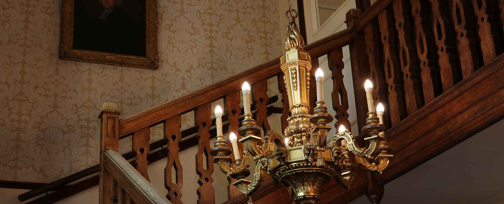 An ornate brass chandelier with lit electric candles mounted on a wall above a wooden staircase, with vintage wallpaper and a framed portrait in the background