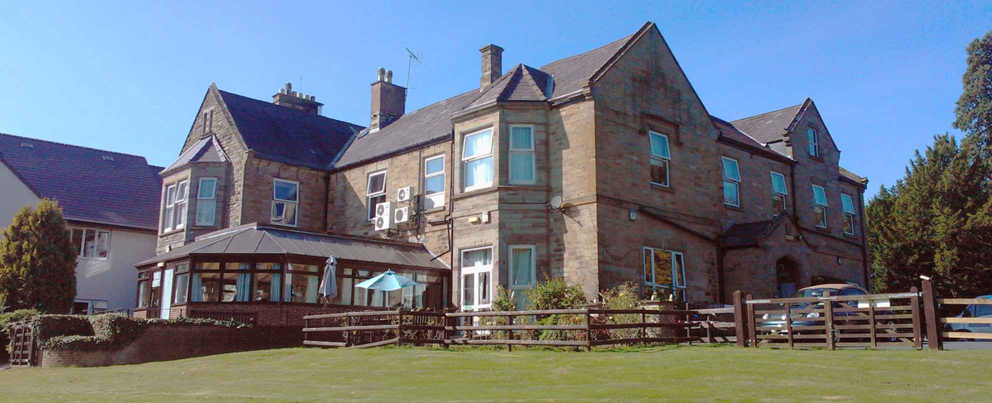 A large two-story stone building with a glass conservatory and a wooden fence in the foreground, set against a clear blue sky