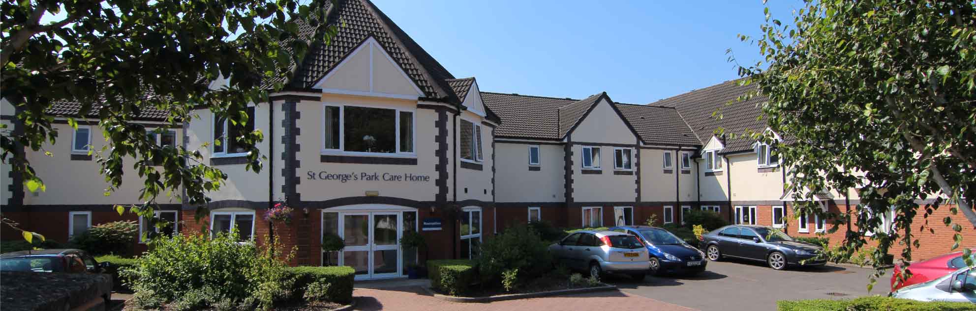 St George's Park front entrance  and car park with several cars parked. The two-story building has white walls with dark roofing, and multiple windows. The care home is flanked by trees on either side. A sign reading “St Georges Park Care Home” is visible above the central entrance.