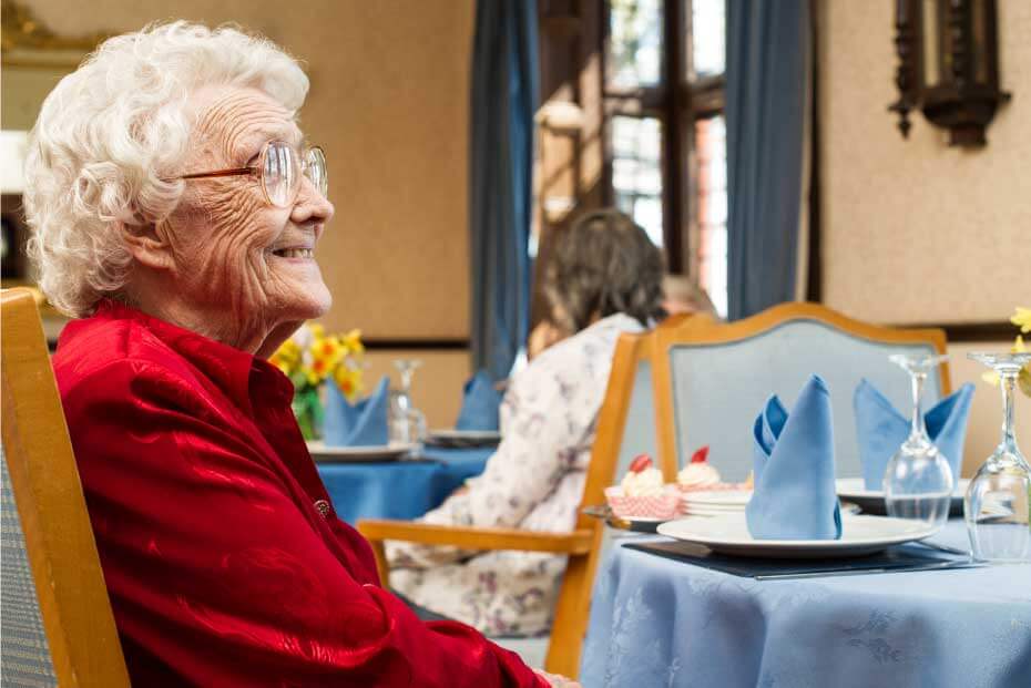 A smiling elderly lady in a red jacket seated at a dining table with a blue tablecloth and plate set,