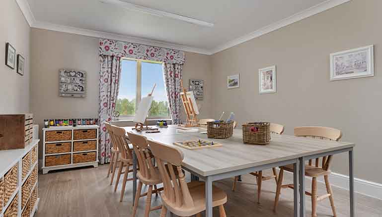 A spacious and organised craftroom with a large table, chairs, storage shelves filled with jars of paint, and an easel positioned by the window.