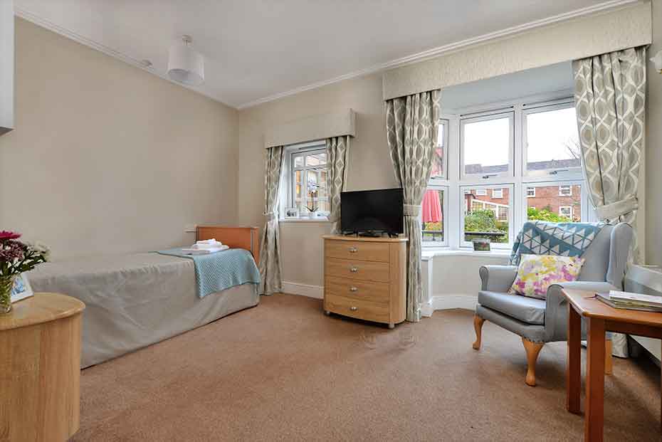 Modern styled bedroom with carpeting,  featuring a single bed, a wooden chest of drawers with a TV on top, a light blue armchair with floral cushions, and patterned curtains.