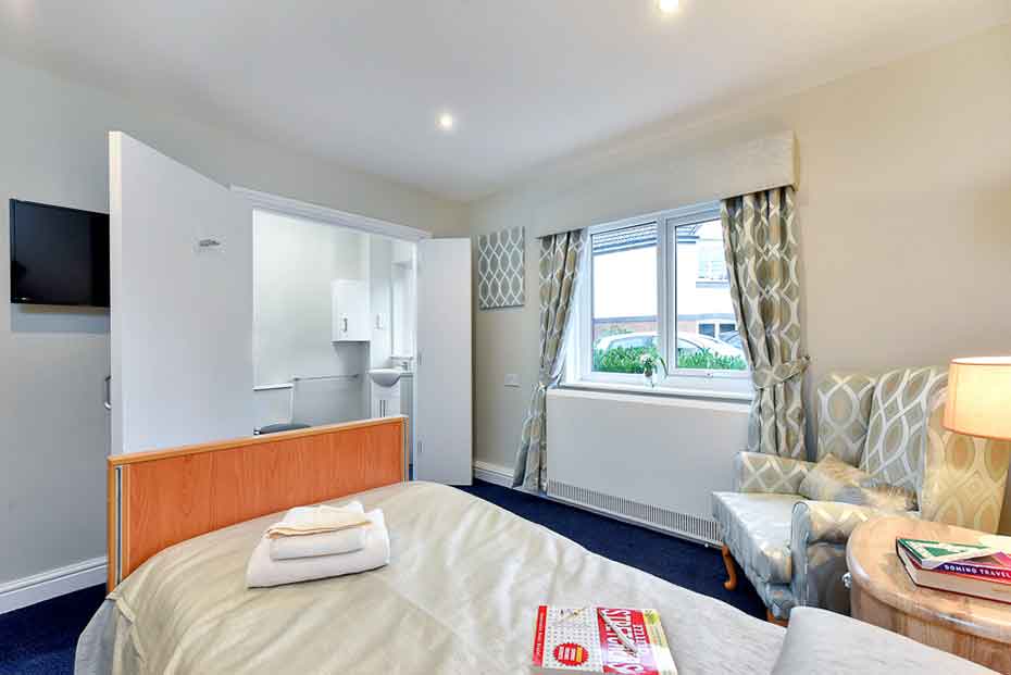 A well-lit bedroom with a double bed adorned with white bedding and a folded towel on it. There is a flat-screen TV on the wall, a small wooden desk with an open magazine, two lamps, and patterned curtains by the window.