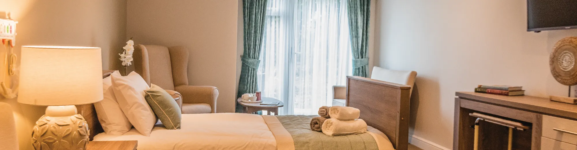 A bedroom with a table lamp, a made bed with rolled towels, an armchair and long green curtains framing a window