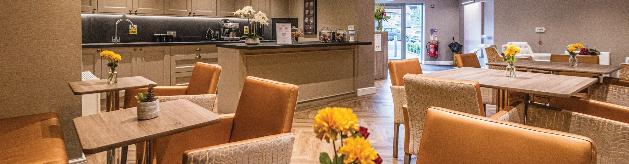 A café area with orange leather chairs, wooden tables topped with yellow flowers and a coffee counter