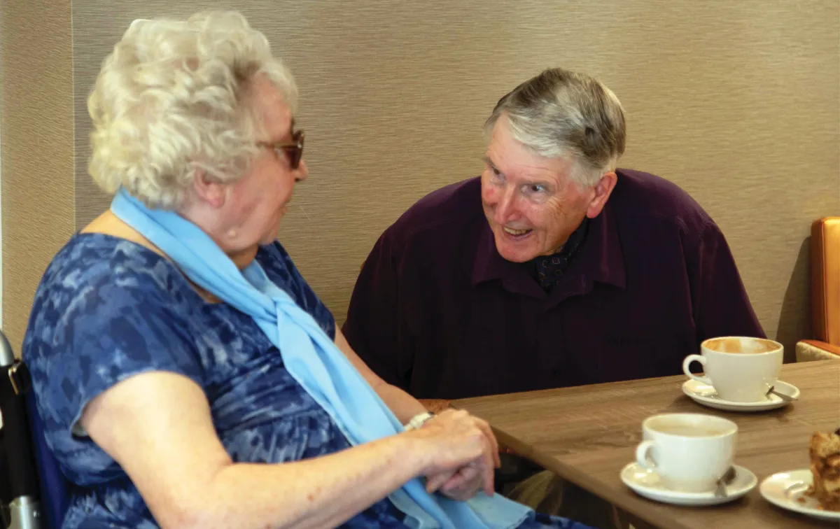 Carole, in a pale blue scarf, chatting with a smiling Dennis across a table set with cups of coffee