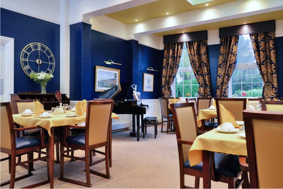 An elegant dining room with multiple set tables, beige chairs, blue walls, and large windows with patterned curtains.