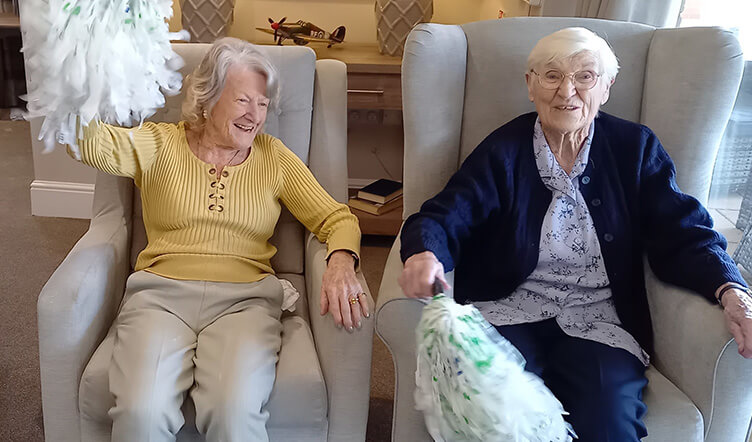 Two elderly ladies seated on armchairs, one in a yellow top and the other in a blue cardigan, using exercise poms poms