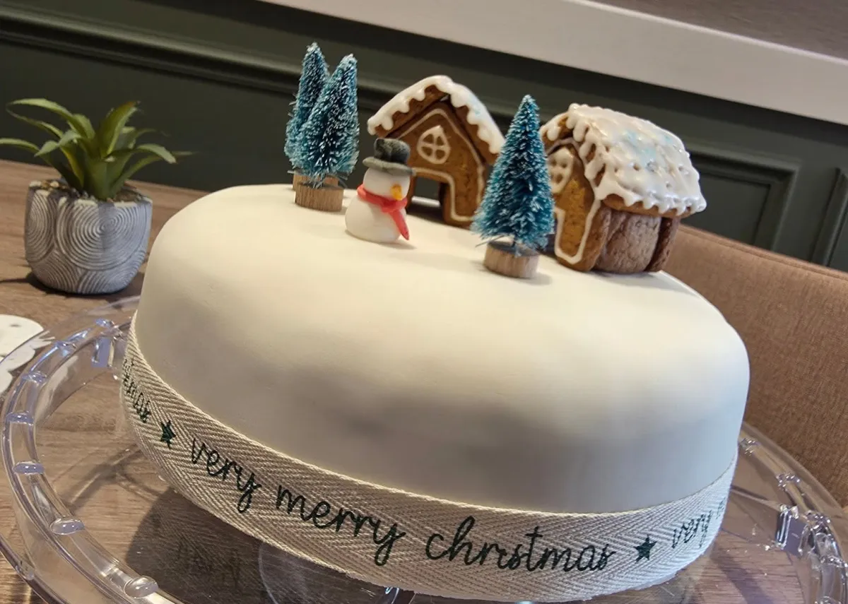 A white iced Christmas cake topped with a snowman, gingerbread houses and miniature trees, wrapped in a “very merry christmas” ribbon