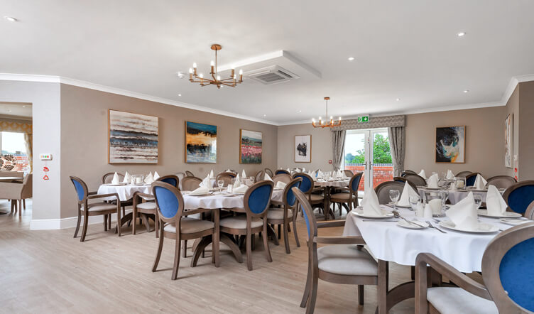 An elegant dining room with multiple set tables, grey and blue upholstered chairs, and paintings on the walls under a coffered ceiling with a chandelier.