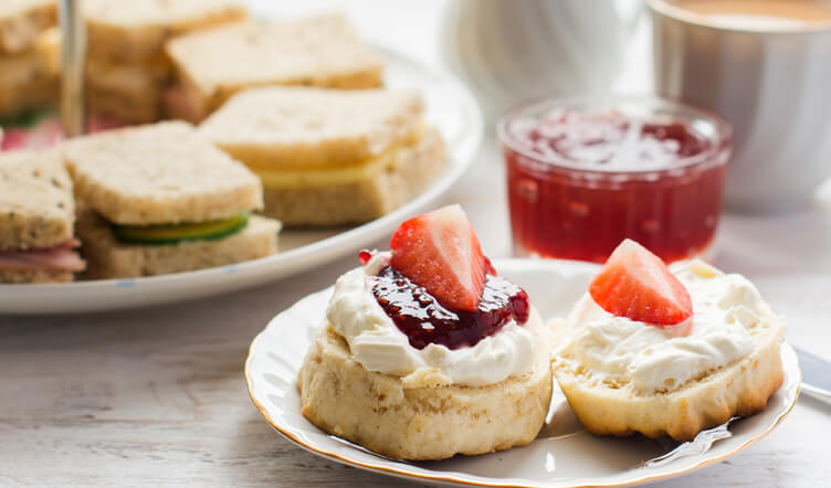 A plate with two scones topped with cream and strawberries, a jar of jam, and sandwiches in the background