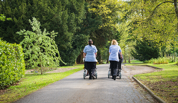 Two healthcare workers pushing wheelchairs on a paved path, with lush green trees and grass on either side