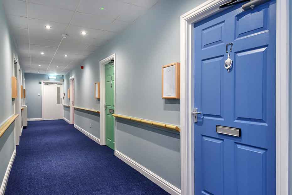 A corridor with blue carpeting and walls painted in light blue and white. There are multiple closed doors with different colours, and a handrail running along one side.