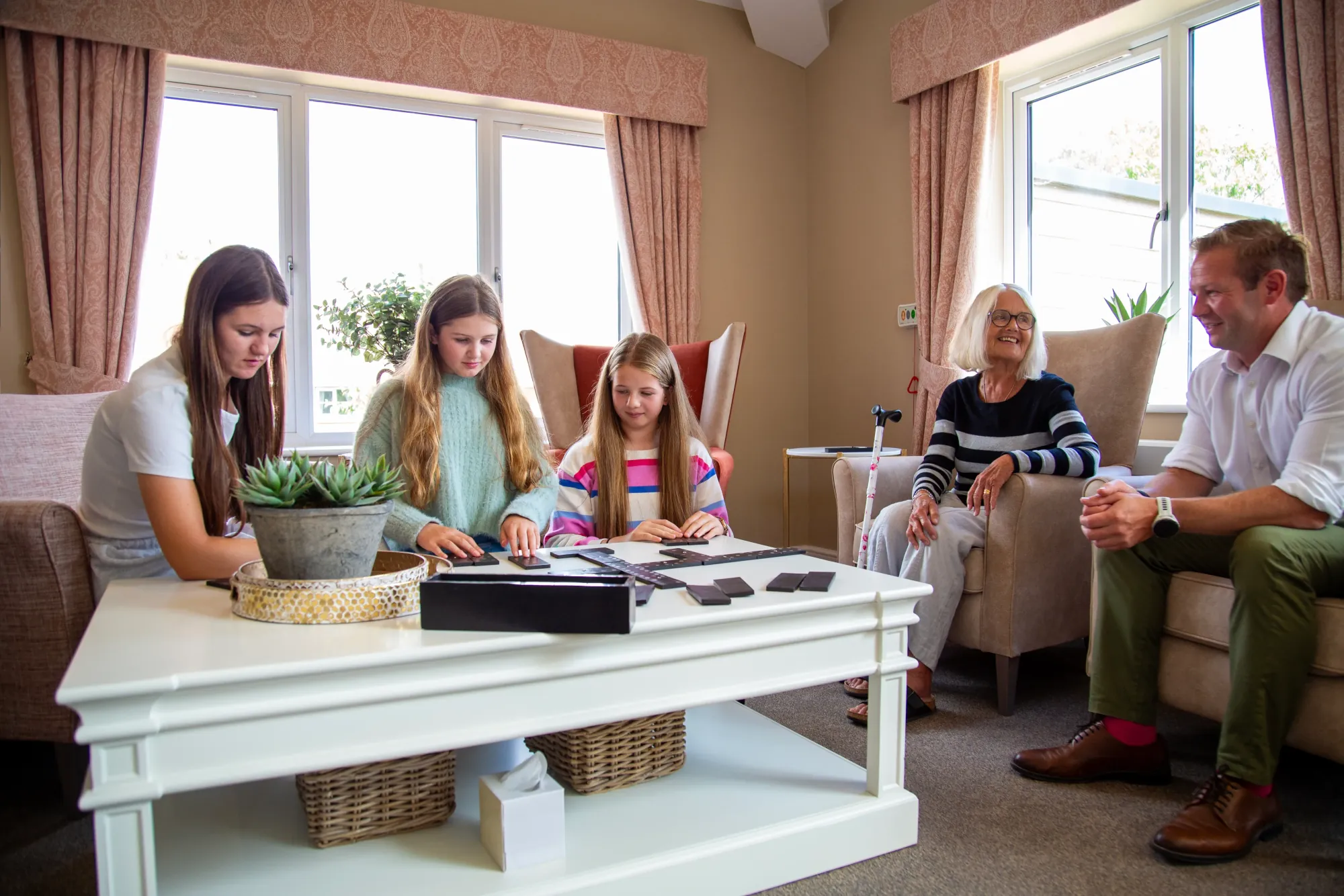A family visiting a resident in a bright lounge at Colwall Care Home, with her granddaughters playing dominoes at the coffee table