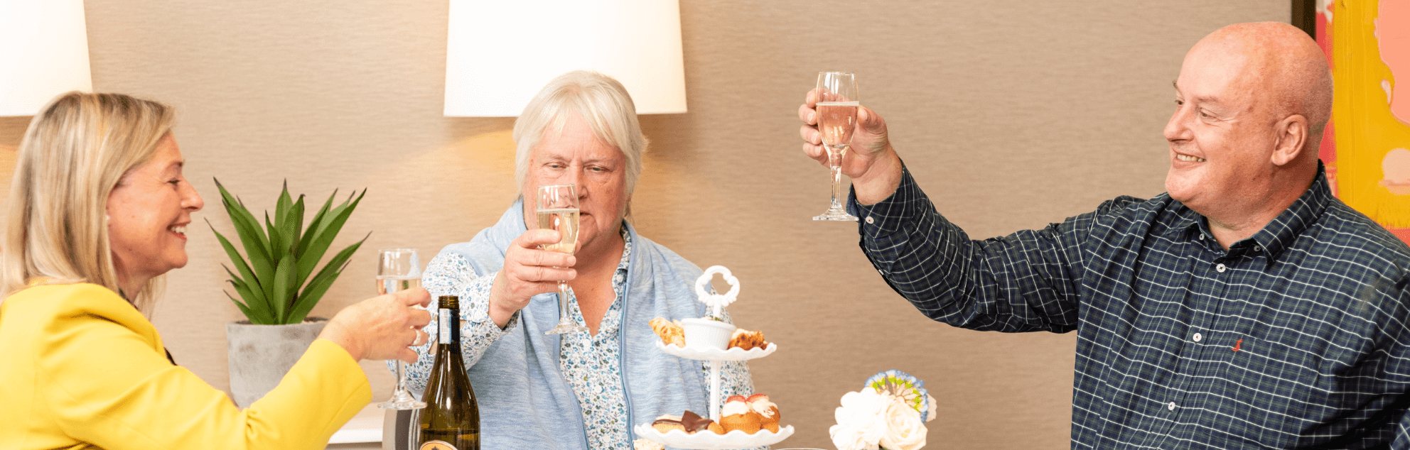 A lady in a yellow jacket raising a champagne flute to a lady in a blue blouse and a man in a grey chequer shirt, with a cake stand in the centre.