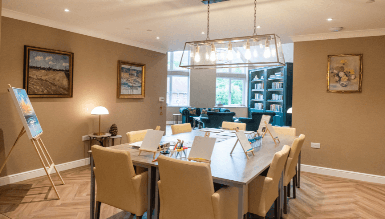 A large room with a large table with mini easels set up, a large handing light above and free standing easel to the side