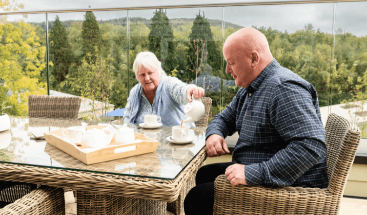 A lady and gentleman sat in wicker chairs pouring cups of tea with the hills in the background