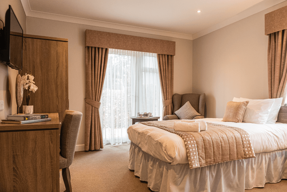 A well-lit, modern bedroom with a large bed, white bedding, a cream throw blanket, , a desk and chair, and a wall-mounted television.