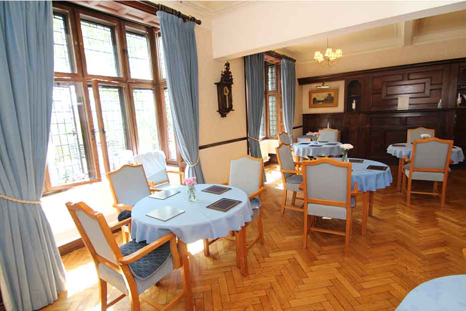 An elegant dining room with tables set with blue tablecloths and brown placemats, wooden flooring, large windows with blue drapes, and a classic wooden sideboard