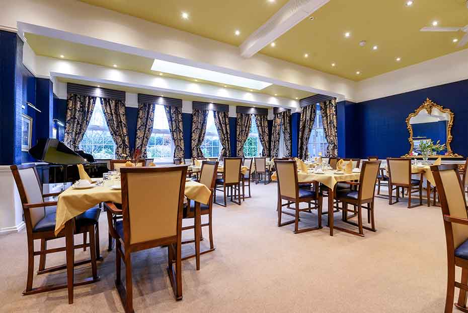 Elegant dining room with multiple set tables, chairs, and a large mirror on the wall. The room has blue walls, patterned curtains, and a gold ceiling with recessed lighting