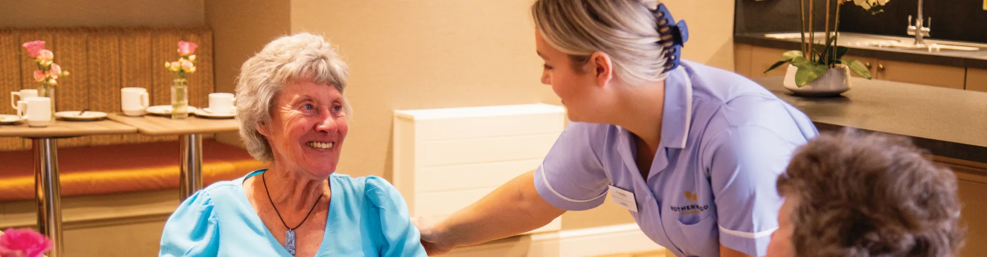 A smiling older woman in a turquoise top looking up at a carer in a lilac uniform who rests a hand on her shoulder