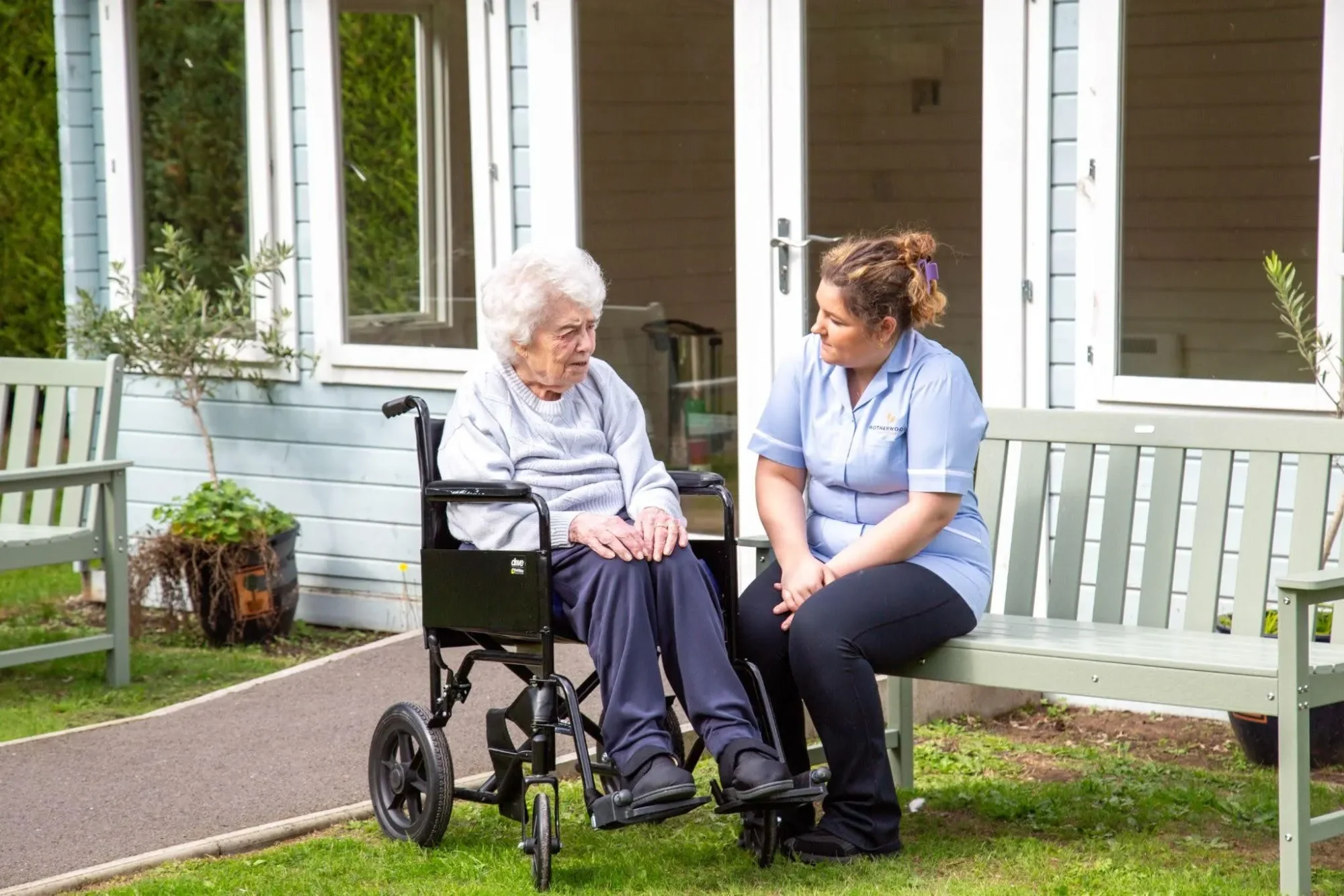A nurse sitting on a garden bench chatting with a resident in a wheelchair at Dorset House