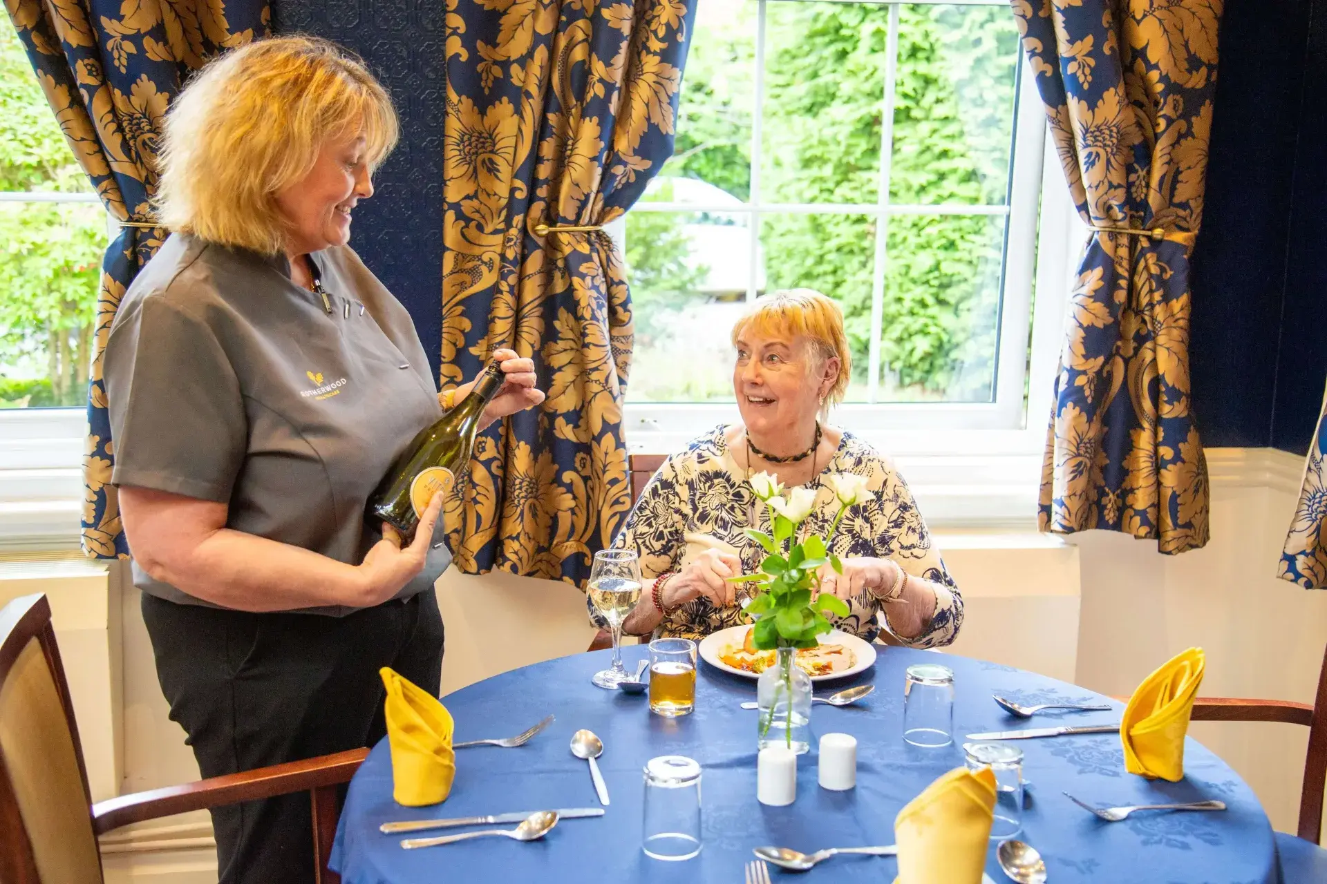 A member of staff showing a bottle of wine to a smiling resident seated at a dining table at Dorset House