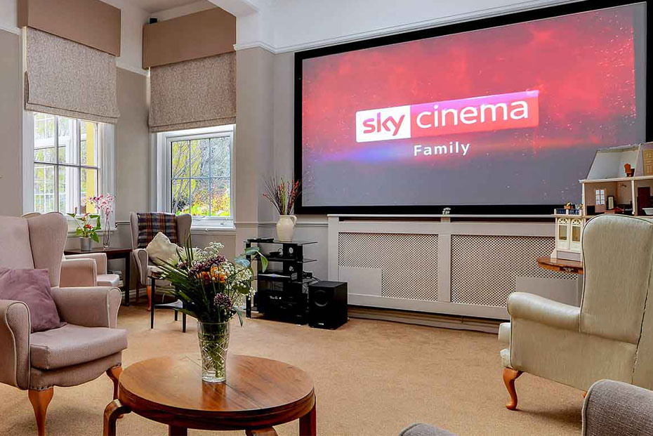 Cinema room with multiple beige armchairs facing a large screen displaying the “Sky Cinema Family” logo. The room has windows on one side, cream walls, and a patterned carpet.