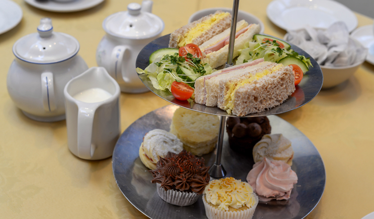 A two-tiered serving tray with an assortment of sandwiches and cakes, accompanied by teapots and a milk jug on a table