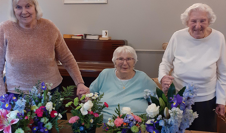 Three elderly ladies, holding hands and standing behind a table adorned with colourful floral arrangements.