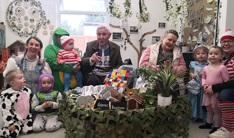 A group of nursery workers and nursery children pose for a group photo, alongside an elderly man and his carer. Several of the adults and children are wearing costumes to commemorate World Book Day
