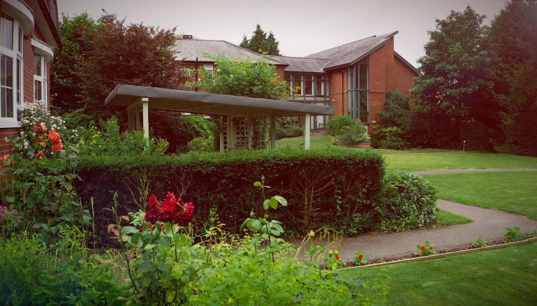 A lush garden leading to a modern building with large windows and a covered walkway, all surrounded by flowering shrubbery