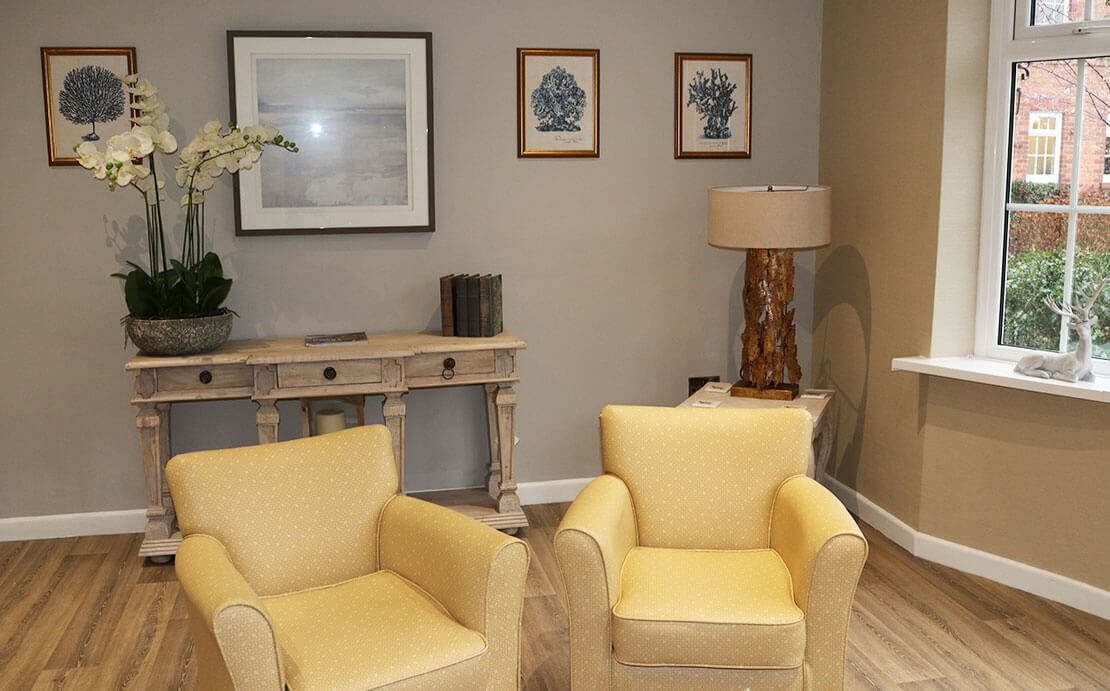 Well-lit room lounge featuring two yellow armchairs, a wooden console table adorned with decorative items, a framed painting on the wall, and three blue botanical prints.