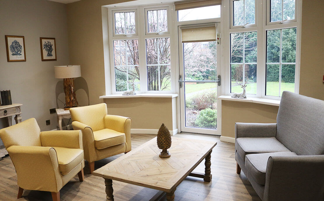 Well-lit living room with two yellow armchairs, a grey sofa, and a wooden coffee table. Large windows in the room provide a beautiful view of an outdoor garden