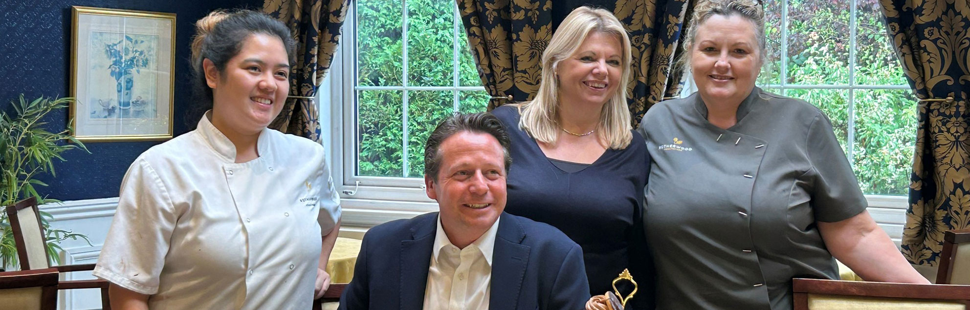 A man and three female healthcare workers smiling and posing for a photo, in front of a window with decorative curtains and furnishings.