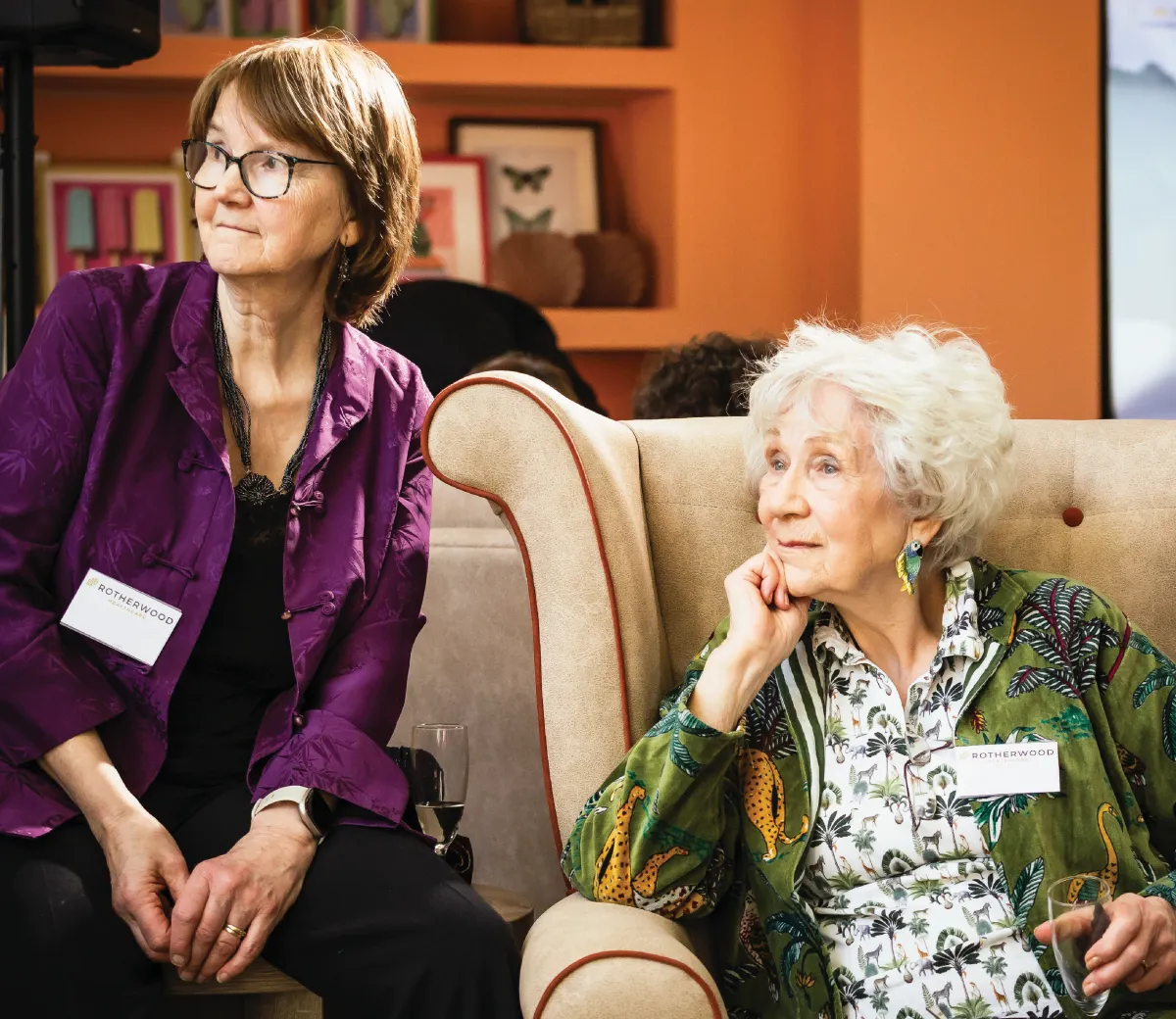 Dr Lynn, in a green jungle-print jacket, sitting in a wingback armchair with her chin resting on her hand, beside a woman in a purple jacket