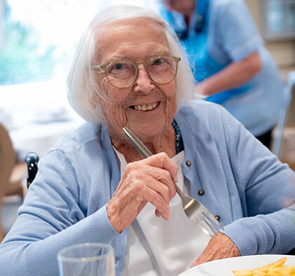 An elderly lady with white hair, wearing a blue cardigan, sits at a table in the dining room and enjoys a meal