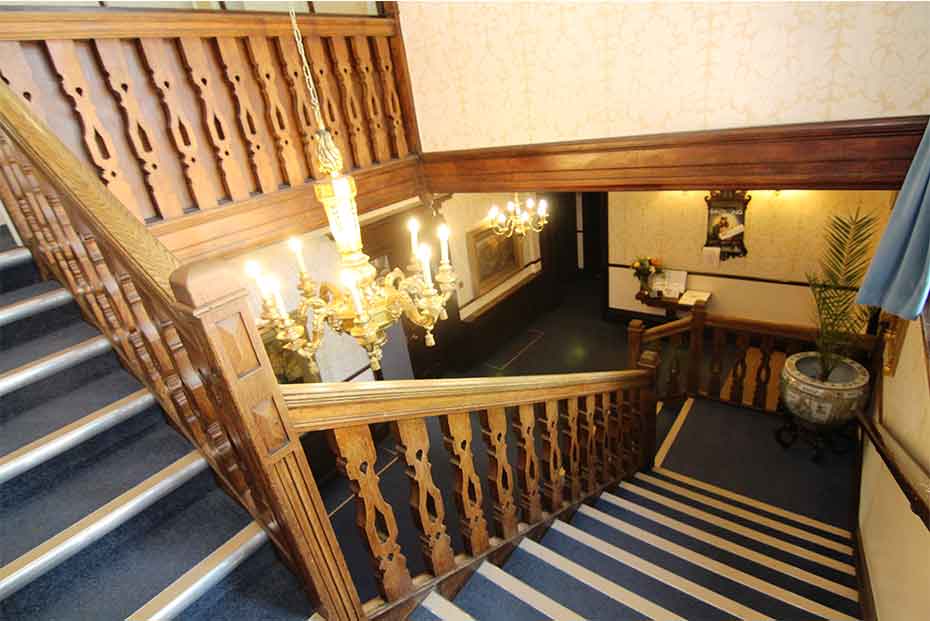 An ornate wooden staircase with decorative spindles, leading to an upper floor in a building with vintage wallpaper and a chandelier.