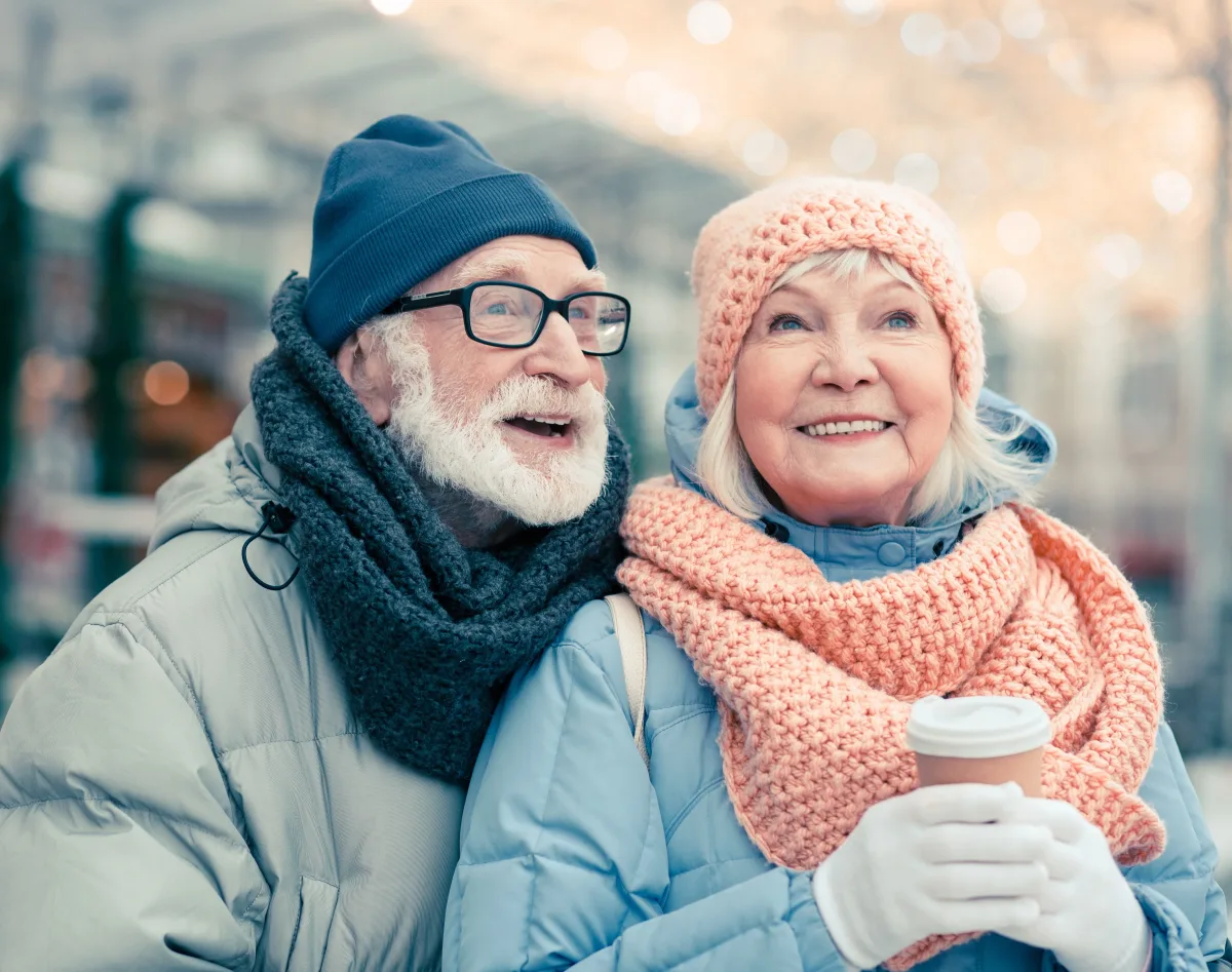 An older couple in woolly hats and scarves smiling outdoors in winter, one holding a takeaway cup