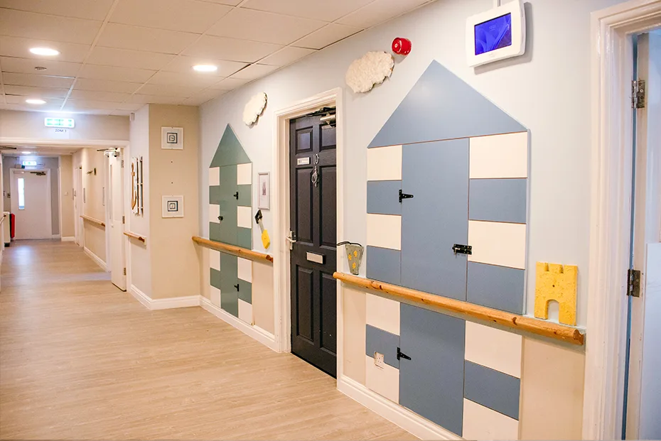 A bright corridor with wood-effect flooring and a wooden handrail, where bedroom doors are framed by blue, white and green beach hut panels with cloud decorations above.