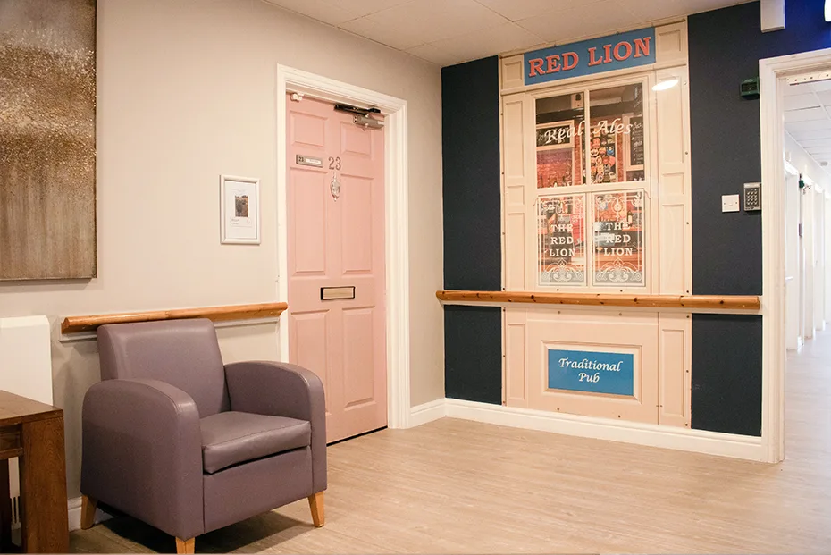 A corridor with a pink bedroom door and a grey armchair, beside a wall dressed as a traditional pub frontage with a 'Red Lion' sign and window.