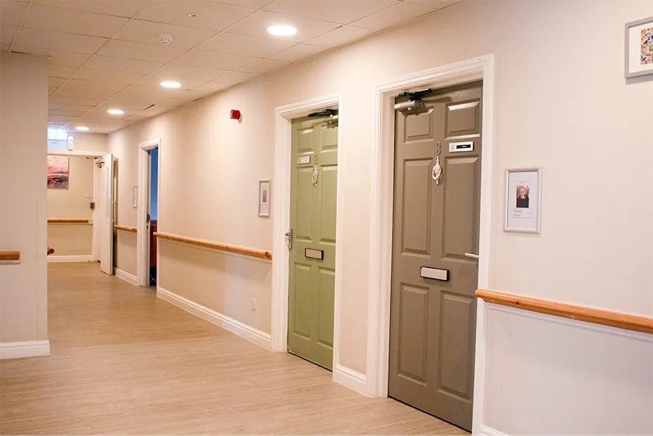 A calm corridor with wood-effect flooring, a wooden handrail, and sage green and grey bedroom doors with framed memory boxes alongside them.